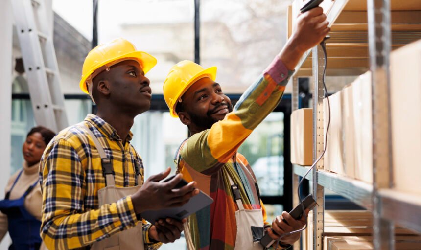 Warehouse smiling colleagues scanning cardboard box barcode and chatting. Two african american post office storehouse workers inspecting customer parcels using scanner tool and tablet